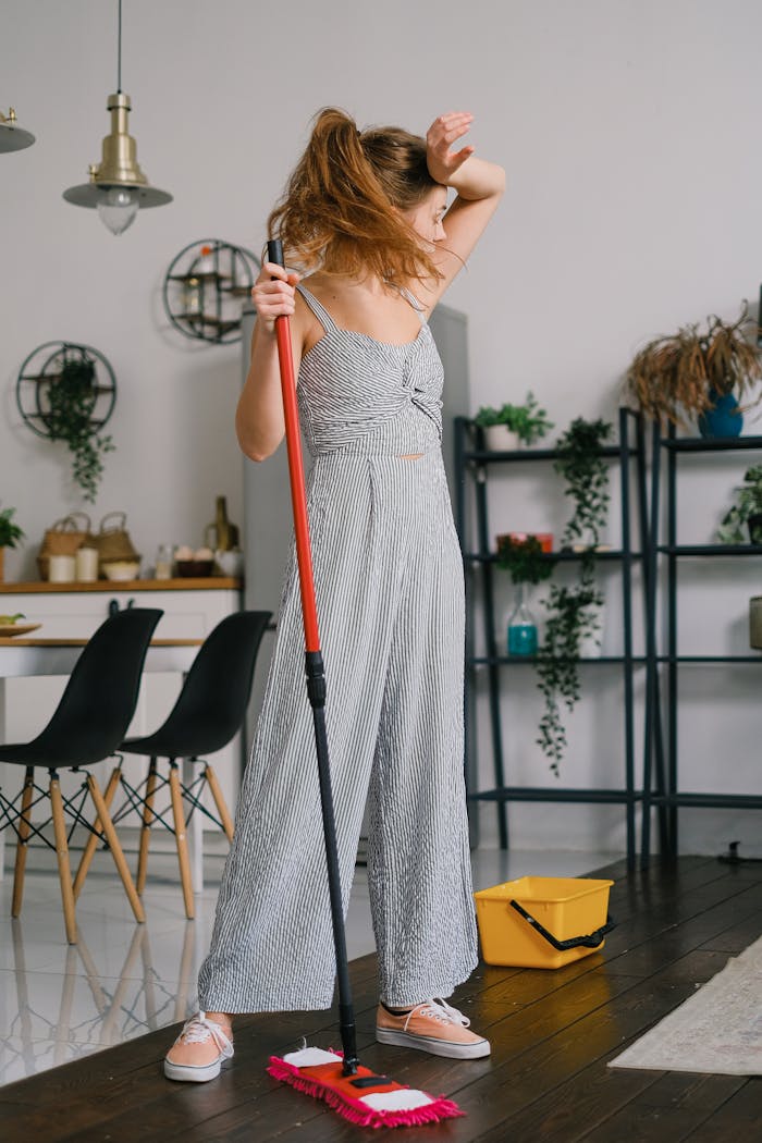A woman in a jumpsuit takes a break from mopping the floor, appearing tired and weary indoors.
